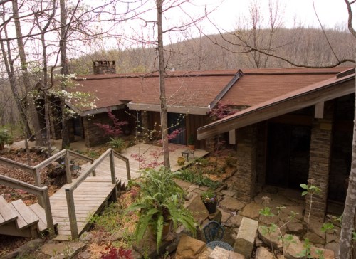 A rustic house surrounded by trees, featuring a wooden deck and stone pathways in a serene, natural setting.