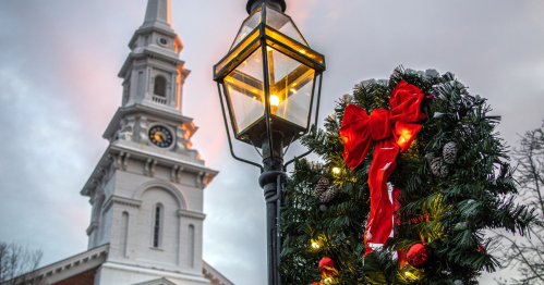 A festive wreath with a red bow and lights hangs on a lamp post, with a church steeple in the background.
