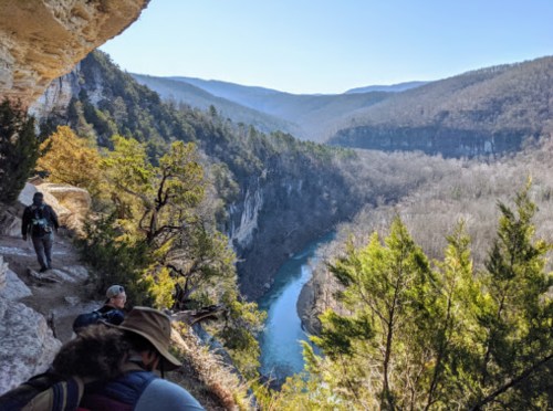 A scenic view of a river winding through a valley, surrounded by cliffs and trees, with hikers in the foreground.