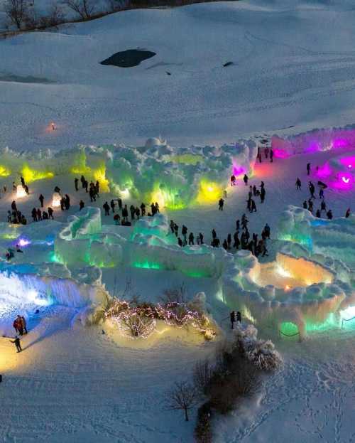 Aerial view of a colorful ice sculpture park with people exploring illuminated ice formations in a snowy landscape.