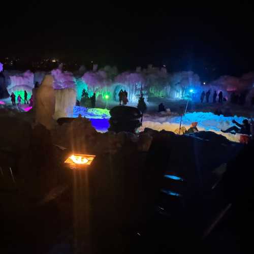 Colorful lights illuminate ice formations and people enjoying a winter night scene. A fire pit glows in the foreground.