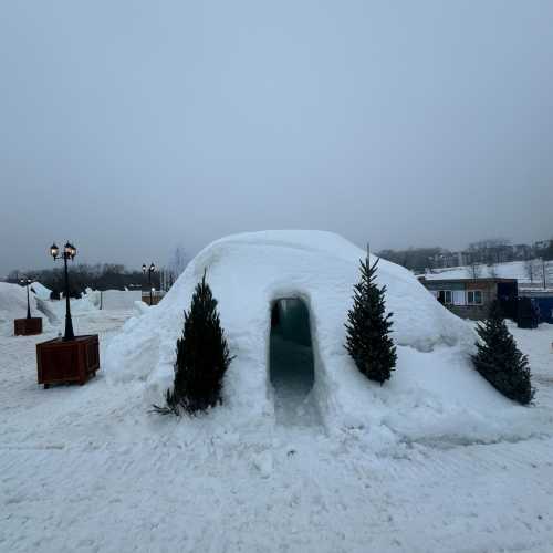 A snow structure resembling an igloo, with a doorway and small evergreen trees on either side, set in a snowy landscape.