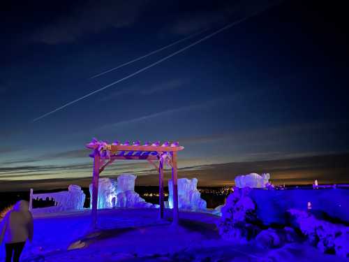 A snowy landscape at night with a lit wooden arch and ice formations, under a starry sky with contrails.