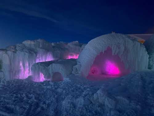 Colorful ice formations illuminated in pink and purple light against a dark sky. Snow covers the ground.