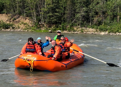 A group of five people in orange life jackets wave from a bright orange raft on a river surrounded by greenery.