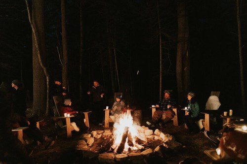 A group of people gathered around a campfire in a dark forest, sitting on wooden chairs and enjoying the warmth.