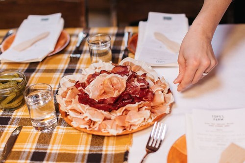 A hand reaches for a platter of assorted cured meats on a checkered tablecloth, with glasses and plates nearby.