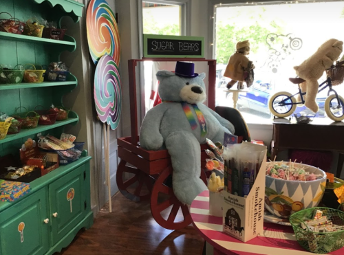 A large blue teddy bear wearing a top hat sits in a cart surrounded by colorful candies and decorations in a cheerful shop.