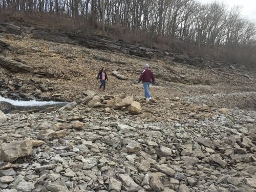Two people walking on a rocky riverbank surrounded by trees in a natural landscape.