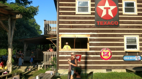 A rustic wooden building with a Texaco sign, people enjoying the outdoors, and a sunny blue sky.