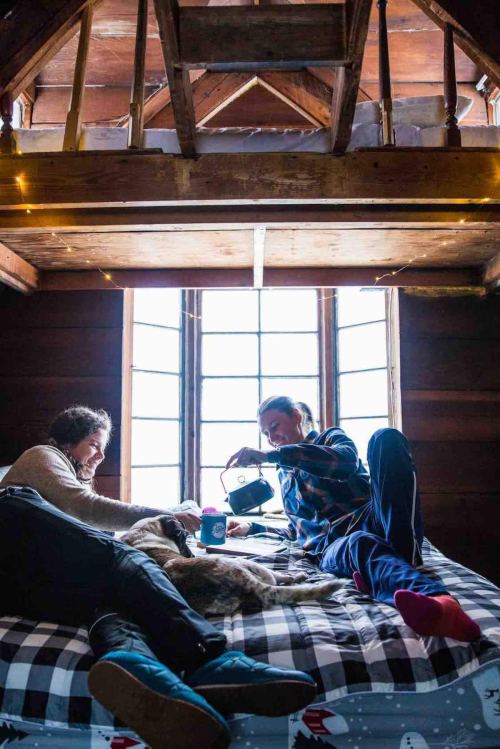 Two people relax on a bed in a cozy cabin, with a dog lying between them and sunlight streaming through the window.