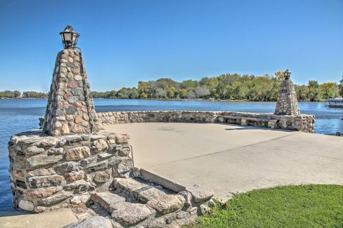 A stone pier with two lamp posts by a calm river, surrounded by trees under a clear blue sky.