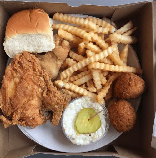 A takeout box with fried chicken, crinkle-cut fries, a roll, coleslaw with a pickle, and two cornbread muffins.