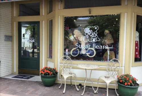 A storefront with a sign reading "Sugar Bears," featuring a teddy bear on a bicycle and outdoor seating with flowers.