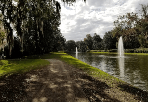 A serene park scene featuring a winding path beside a pond with fountains and lush greenery under a cloudy sky.