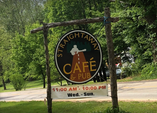 Sign for "Straightaway Café & Pub" with operating hours, surrounded by greenery. Open Wednesday to Sunday, 11 AM - 10 PM.