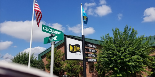 A street sign for Cucumber and Vine, with a Mr. Olive restaurant sign and flags in the background under a blue sky.