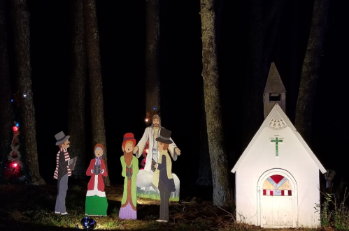 A festive scene with cutout figures of carolers and a church, set in a dark forest with holiday lights.