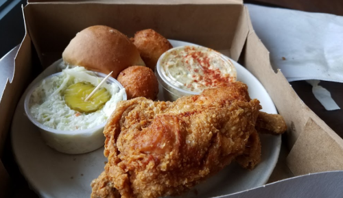 A takeout box with fried chicken, coleslaw, a roll, and two hushpuppies.