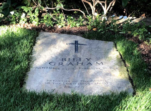 Gravestone of Billy Graham, inscribed with his name, dates, and a biblical verse, surrounded by grass and flowers.