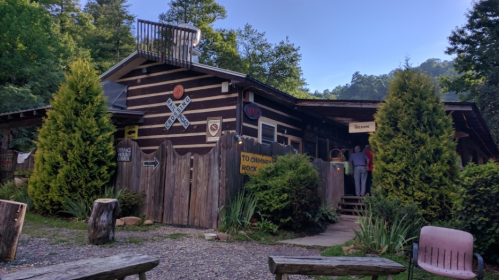 A rustic log cabin surrounded by greenery, featuring signs and a welcoming entrance.