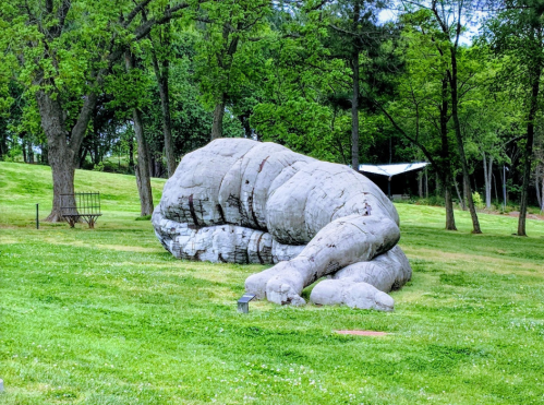 A large, abstract sculpture resembling a hand resting on the grass, surrounded by trees in a park setting.
