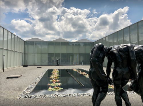 A serene courtyard with a reflective pond, surrounded by modern glass architecture and sculptures under a cloudy sky.