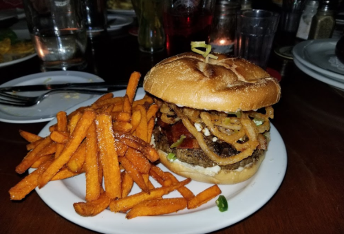 A juicy burger topped with crispy onions and bacon, served with a side of sweet potato fries on a white plate.