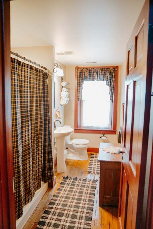 A cozy bathroom with a shower curtain, window, toilet, and a rug, featuring warm colors and rustic decor.