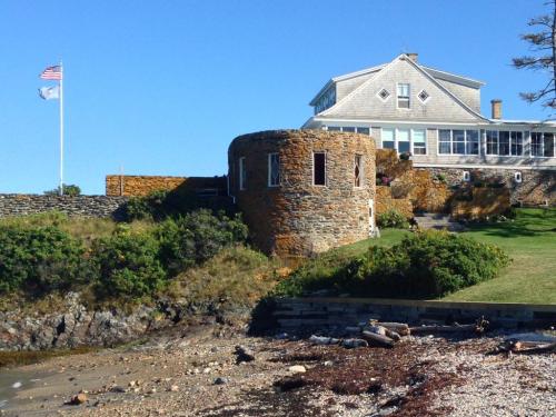 A coastal scene featuring a stone round building, a house, and flags against a clear blue sky.