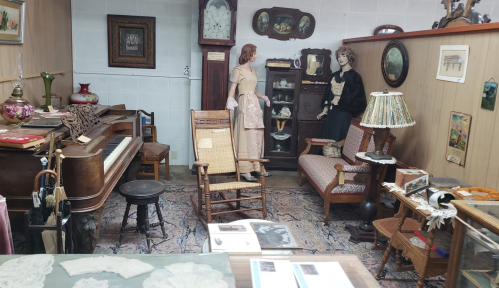 A vintage room display featuring a piano, rocking chair, antique furniture, and two mannequins in period clothing.