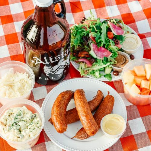 A picnic spread featuring sausages, a fresh salad, sides, and a large brew jug on a red checkered tablecloth.