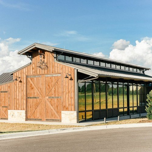 A modern wooden barn with large glass windows, set against a blue sky with fluffy clouds.