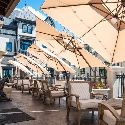 A sunny outdoor patio with wooden chairs and large beige umbrellas, surrounded by a stylish building.