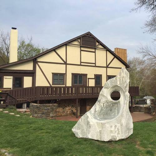 A large, abstract stone sculpture sits in front of a rustic, two-story house with a wooden deck.