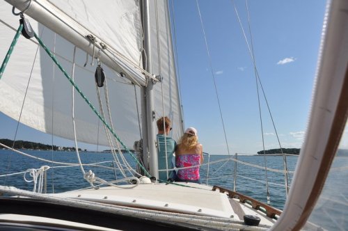 Two people sit on a sailboat, enjoying a sunny day on the water with sails raised and a clear blue sky.