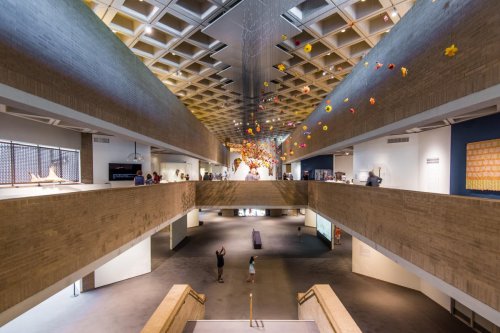 A spacious art gallery with a modern design, featuring colorful hanging decorations and visitors exploring the space.