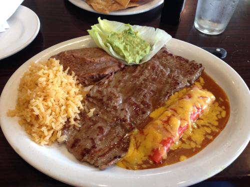 A plate of grilled steak with rice, refried beans, enchiladas topped with cheese, and lettuce with guacamole.