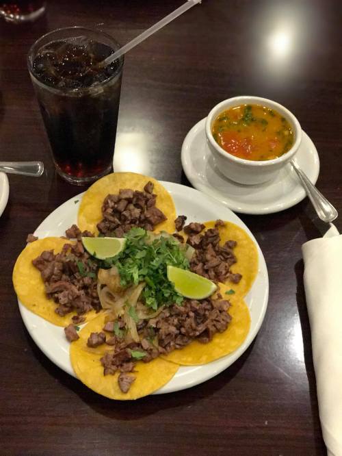 A plate of tacos filled with beef, topped with onions and cilantro, served with lime wedges and a bowl of soup.