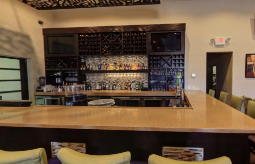 A modern bar with a sleek countertop, shelves of liquor, and a stylish backdrop in a well-lit space.
