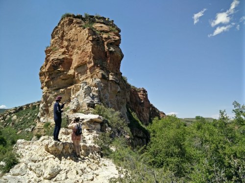 Two people explore a rocky outcrop surrounded by greenery under a clear blue sky.