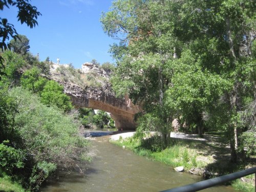 A stone bridge arches over a river, surrounded by lush greenery and rocky cliffs under a clear blue sky.