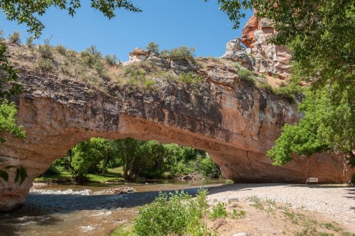 A natural stone arch spans over a river, surrounded by lush greenery and rocky cliffs under a clear blue sky.