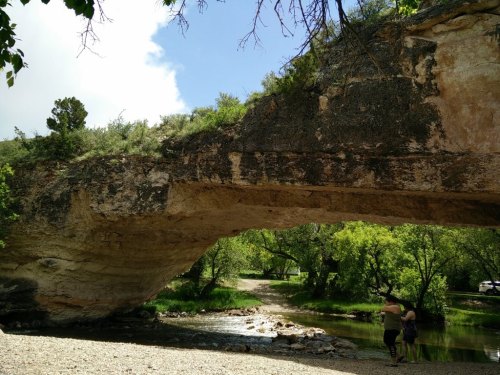 A natural stone arch over a river, with two people standing nearby in a lush green landscape.