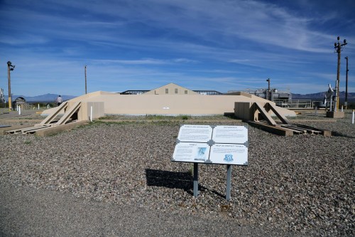 A barren landscape featuring a low, beige structure and informational signs on gravel under a clear blue sky.