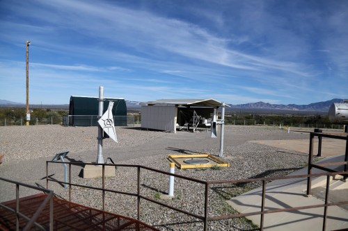 A rural landscape featuring a small shed, equipment, and a clear blue sky with distant mountains.