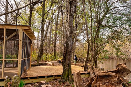 A wooden deck by a river, surrounded by trees and greenery, with a person standing in the background.