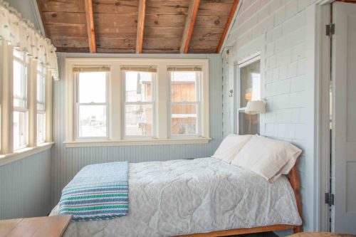 Cozy bedroom with a wooden bed, light blue walls, and large windows letting in natural light.