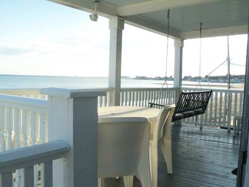 A beachside porch with a swing and table, overlooking calm waters and a distant shoreline.