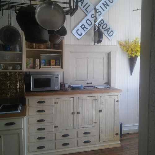 A rustic kitchen with a wooden cabinet, microwave, and decorative wall signs, featuring yellow flowers in a wall vase.